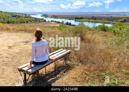 A young woman sits on a wooden bench and looks at the calm Southern Bug River on a sunny autumn day. Stock Photo
