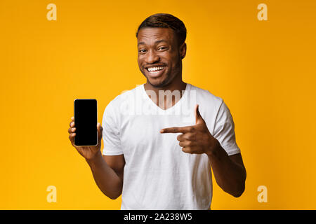 African American Man Holding Smartphone Pointing Finger At Empty Screen Standing On Yellow Studio Background. Check This Out. Mockup Stock Photo