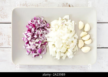 Organic diced red and white onion and garlic cloves on beige plate on weathered white board background.  Vegetable preparation for cooking. Stock Photo