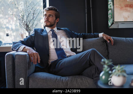 Young and charming businessman is thinking about business while sitting on the sofa in his modern office. Stock Photo