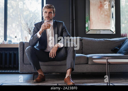 Young and charming businessman is thinking about business while sitting on the sofa in his modern office. Stock Photo