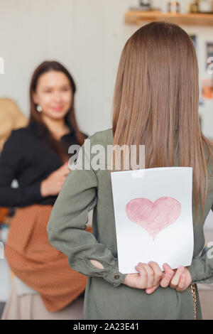 Girl holds a card made for mom on holiday behind her back. Stock Photo