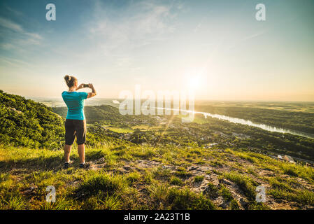 A Girl Standing on the Edge of the Cliff and Taking Photograph of the Sunset and the Valley with the River Stock Photo