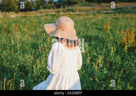 Beautiful romantic preteen girl in white dress and straw hat from behind at the meadow, golden hour Stock Photo