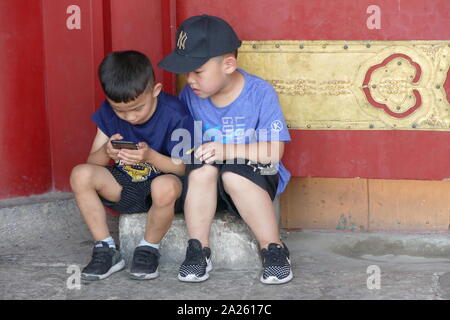 Two small boys play with a mobile phone at the Red gilded gate to the Forbidden City, Beijing, China. The Forbidden City is a palace complex in central Beijing, China. It houses the Palace Museum, and was the former Chinese imperial palace from the Ming dynasty to the end of the Qing dynasty (the years 1420 to 1912). The Forbidden City served as the home of emperors and their households and was the ceremonial and political center of Chinese government for almost 500 years. Stock Photo