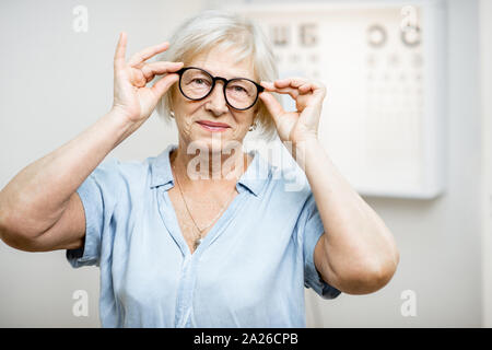 Portrait of a happy senior woman wearing eyeglasses in front of eye chart in ophthalmology office. Concept of checking eyesight and selecting glasses Stock Photo
