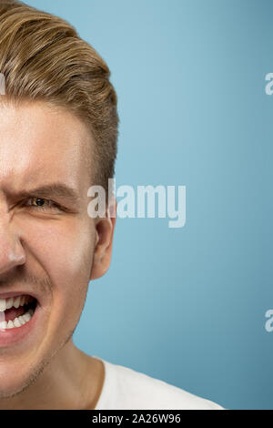 Caucasian young man's close up portrait on blue studio background. Beautiful male model with well-kept skin. Concept of human emotions, facial expression, sales, ad, beauty. Angry screaming. Stock Photo