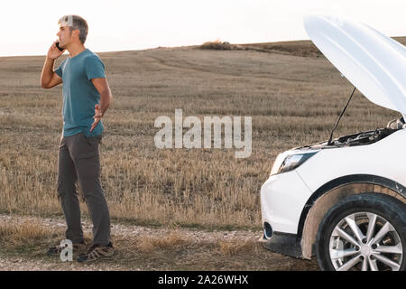 An angry man in the middle of the field calling the insurance of his broken down car for assistance at sunset. Lifestyle concept Stock Photo