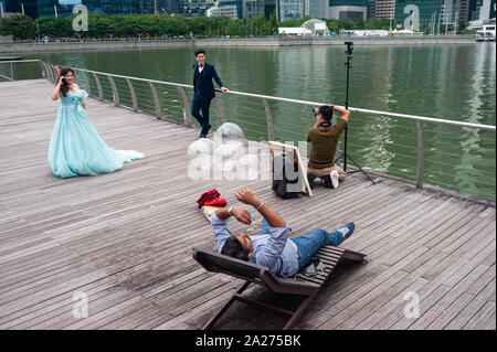 10.05.2019, Singapore, , Singapore - A man relaxes on a couch on the promenade in Marina Bay, while a photographer takes wedding photos in the backgro Stock Photo