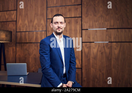 Photo of young man standing near table with laptop on background of wooden wall in office Stock Photo