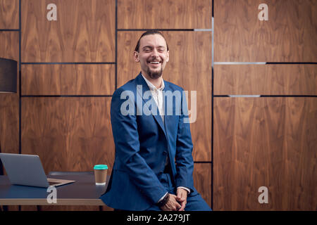 Photo of happy businessman standing near table with laptop in office Stock Photo