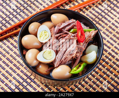 close-up of Jangjorim, fall apart beef braised in soy sauce with quail eggs, garlic cloves and chili pepper slices served in a black bowl on a rustic Stock Photo