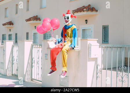 a creepy clown wearing a colorful yellow, red and blue costume, holding a bunch of red balloons in his hand, sitting in front of a row of houses Stock Photo