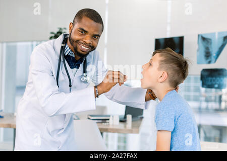 Happy African American male doctor watching patient boy with sore throat in clinic. The doctor examining little boy's throat at hospital Stock Photo