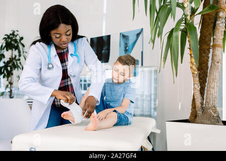 Putting bandage. Dark-skinned woman pediatrician wearing white coat putting bandage on leg of little boy Stock Photo