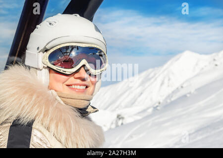 Portrait of young adult beautiful happy caucasian woman smiling on ski-lift at alpine winter skiing resort. Girl in fashion ski suit, goggles and Stock Photo