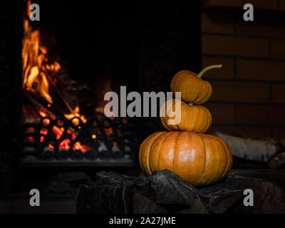 Three halloween pumpkins stand on top of each other on birch firewood. In the background, flames burst from the fireplace. Mystical holiday Halloween. Stock Photo