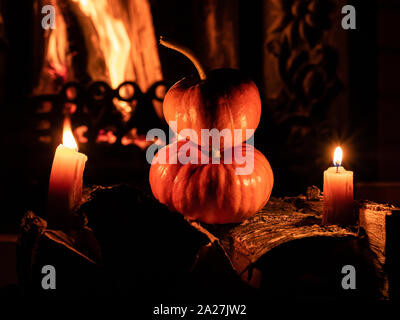 Two halloween pumpkins standing in the dark on birch logs surrounded by burning candles. In the background is a flame from a fireplace. Mystical holid Stock Photo