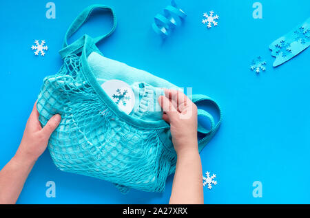 Packing presents, blue sweater  in string or mesh bag. Wintertime composition, flat lay on blue paper with serpantine and snowflakes. Christmas gifts Stock Photo