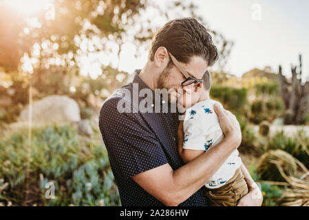 Portrait of father embracing young toddler son with sun behind them Stock Photo