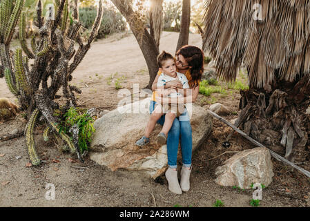 Mother embracing and kissing young son on rock in sunny cactus garden Stock Photo
