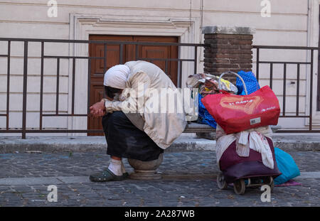 Seated homeless woman with bowed covered head with all her belongings on a trolley behind her in plastic bags. Stock Photo