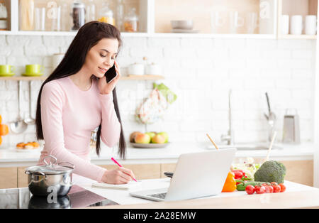 Young woman working at home, talking on phone and taking notes Stock Photo