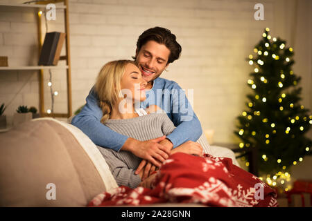 Loving couple embracing near Christmas tree, resting on sofa Stock Photo