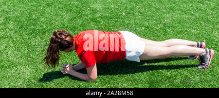 A high school girl is working on her core by performing a front plank exercise on green turf during track and cross country practice. Stock Photo