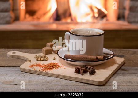 Indian traditional Masala chai tea in a mug and kitchen herbs, over cozy fireplace background with copy space for text. Stock Photo
