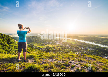 A Girl Standing on the Edge of the Cliff and Taking Photograph of the Sunset and the Valley with the River Stock Photo