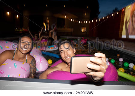 Happy, carefree young couple with inflatable rings taking selfie with camera phone in summer swimming pool