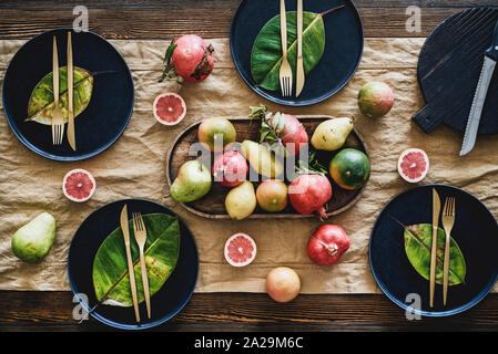 Fall or Autumn table styling for holiday dinner. Flat-lay of black dinnerware with fruit and fallen leaves for decoration over wooden table, top view. Stock Photo