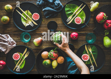 Autumn table styling for holiday dinner. Flat-lay of black dinnerware with fruit and leaves and female hands holding jug over rustic wooden table, top Stock Photo