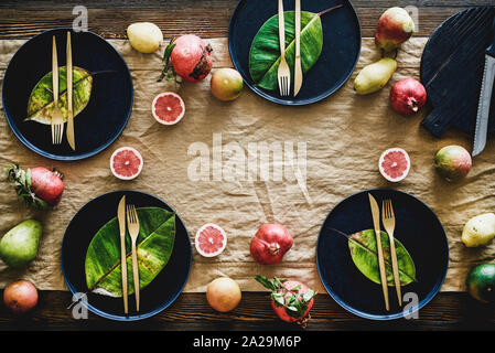 Autumn table styling for holiday dinner. Flat-lay of black dinnerware with fruit and fallen leaves for decoration over wooden table, top view, copy sp Stock Photo