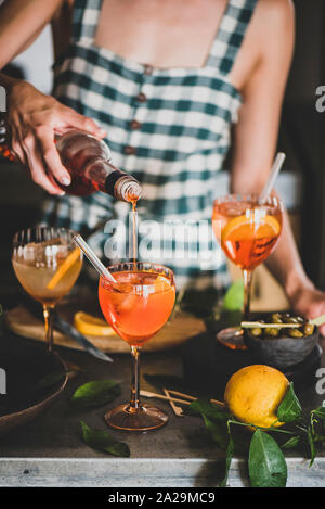 Young woman in checkered dress pouring Aperol to Aperol Spritz aperitif drink with orange in kitchen. Summer refreshing drink concept Stock Photo