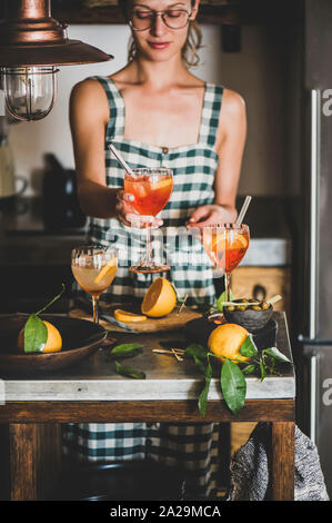 Young smiling woman in checkered dress holding Aperol Spritz aperitif drink with orange in hands, kitchen at background. Summer refreshing drink conce Stock Photo