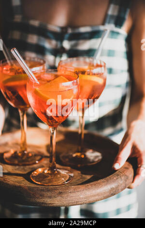 Young woman in checkered dress holding tray with glasses of Aperol Spritz aperitif cold drink with orange in glass, selective focus. Summer refreshing Stock Photo