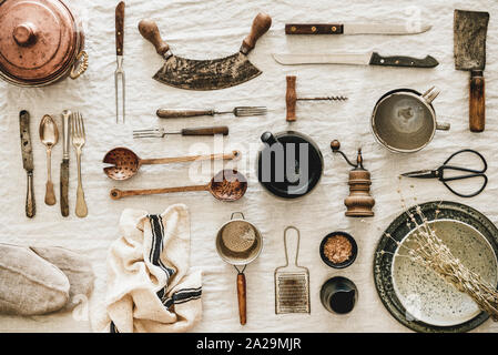 Flat-lay of various kitchen utensils, rustic tablewear, plates, dishes, glasswear, pan, scissors, corkscrew, textile for cooking over white linen tabl Stock Photo
