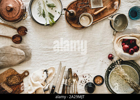 Flat-lay of various kitchen utensils, rustic tablewear, plates, dishes, glasswear, pan, mitten, textile for cooking over white linen tablecloth backgr Stock Photo