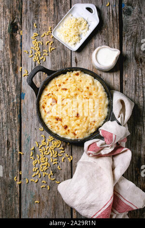 Classic american dish baked mac and cheese in cast iron pan with kitchen towel and ingredients above over old wooden background. Flat lay, space Stock Photo