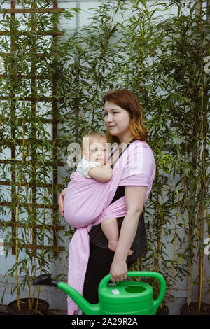 Young redhead babywearing mother carry her sleepy one year old baby girl in a pink wrap sling while gardening Stock Photo