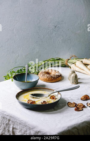 Parsnip cream soup in ceramic bowl with butter sauce, sun dried pears, bundle of fresh parsnip, bagel bread and herbs on kitchen table with white tabl Stock Photo