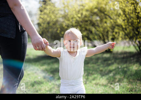 One year old taking first steps, holding mother's hand on a sunny spring day in a park Stock Photo