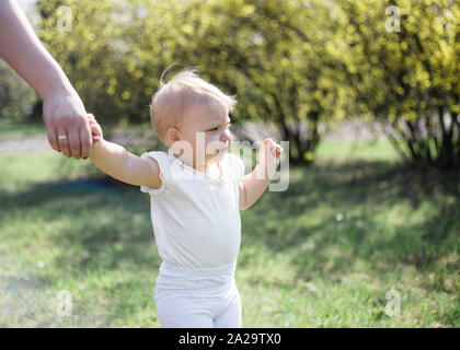 One year old taking first steps, holding mother's hand on a sunny spring day in a park Stock Photo