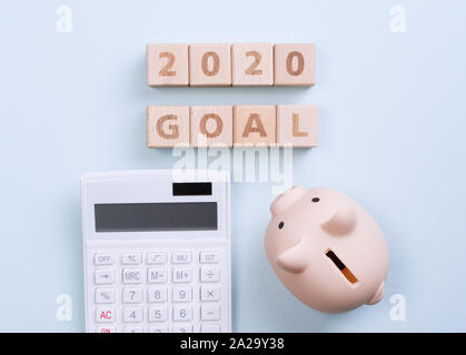 Abstract 2020 financial goal design concept - geometric wood blocks cubes on blue table background with piggy bank, top view, flat lay, copy space. Stock Photo