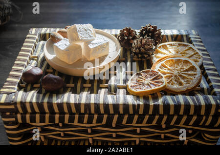 Delicious white Turkish delight on a wooden plate sprinkled with powdered sugar. Oriental pieces of sweets next to cones, chestnuts and dried orange r Stock Photo