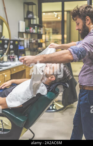 Barber covering client with towel before giving him a shave at barbershop. Stock Photo