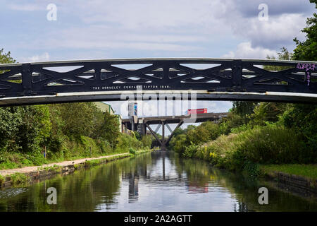 View along the main line canal into the centre of Birmingham with a traditional iron bridge in the foreground and the M5 motorway behind Stock Photo