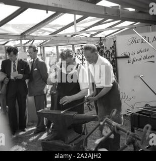 1950s, historical, two blacksmiths at work at an anvil on a stall inside a roofed enclosure at an agricultural show, promoting rural skills, Sussex, England, UK. A blacksmith, also known as a farrier or smithy is a person skilled at metalwork. Stock Photo